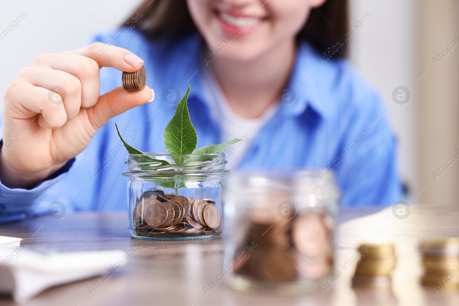 Photo of Woman putting coins into glass jar at wooden table indoors, closeup