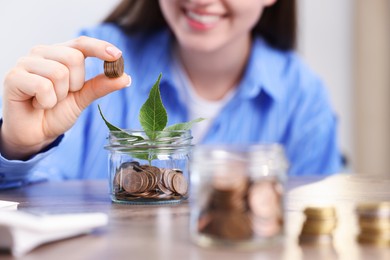 Photo of Woman putting coins into glass jar at wooden table indoors, closeup