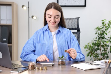 Photo of Woman putting coin into glass jar and using calculator at wooden table indoors