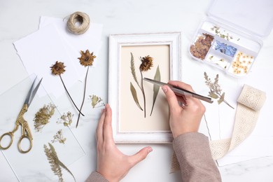 Photo of Woman putting dry flowers into picture frame at white marble table, top view