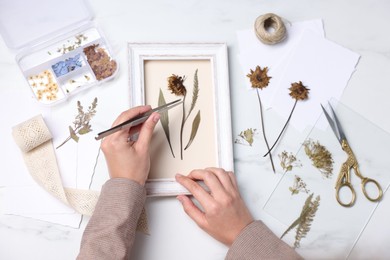 Photo of Woman putting dry flowers into picture frame at white marble table, top view