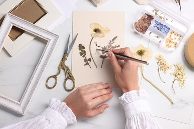 Photo of Woman putting dry flowers onto paper sheet at white marble table, top view