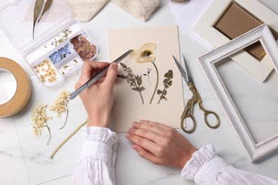 Photo of Woman putting dry flowers onto paper sheet at white marble table, top view