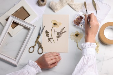 Photo of Woman putting dry flowers onto paper sheet at white marble table, top view