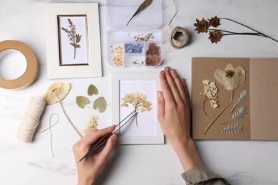 Photo of Woman putting dry flowers into picture frame at white marble table, top view