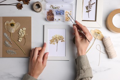 Photo of Woman putting dry flowers into picture frame at white marble table, top view