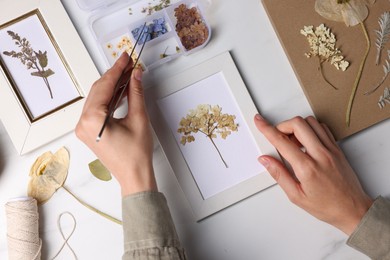 Photo of Woman putting dry flowers into picture frame at white marble table, top view