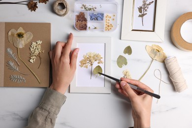 Photo of Woman putting dry flowers into picture frame at white marble table, top view