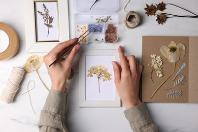 Photo of Woman putting dry flowers into picture frame at white marble table, top view