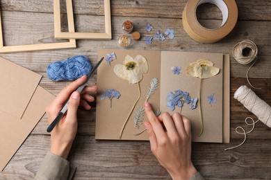Photo of Woman putting dry flowers into notebook at wooden table, top view