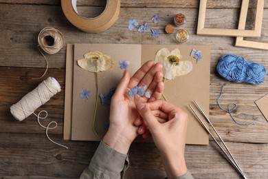 Photo of Woman putting dry flowers into notebook at wooden table, top view