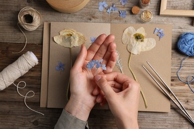 Photo of Woman putting dry flowers into notebook at wooden table, top view