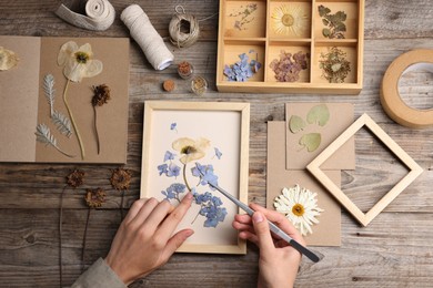 Photo of Woman putting dry flowers into picture frame at wooden table, top view
