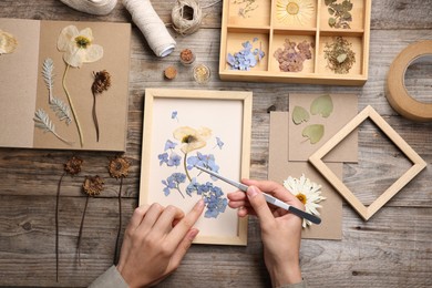 Photo of Woman putting dry flowers into picture frame at wooden table, top view