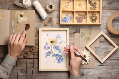 Photo of Woman putting dry flowers into picture frame at wooden table, top view