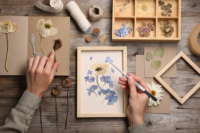 Photo of Woman putting dry flowers into picture frame at wooden table, top view