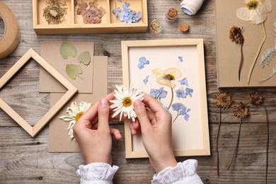 Photo of Woman putting dry flowers into picture frame at wooden table, top view