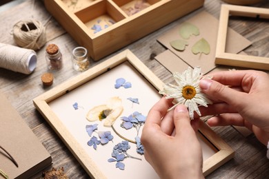 Photo of Woman putting dry flowers into picture frame at wooden table, closeup
