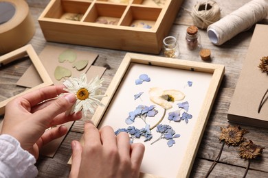 Photo of Woman putting dry flowers into picture frame at wooden table, closeup