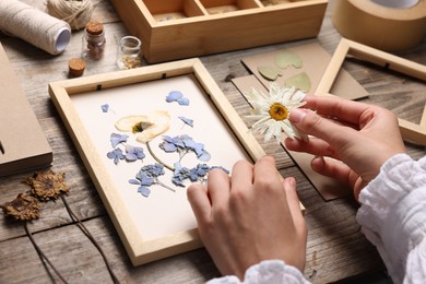 Photo of Woman putting dry flowers into picture frame at wooden table, closeup