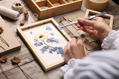 Photo of Woman putting dry flowers into picture frame at wooden table, closeup