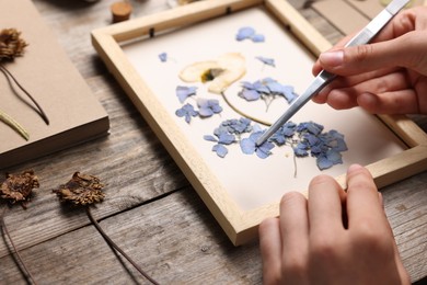 Photo of Woman putting dry flowers into picture frame at wooden table, closeup