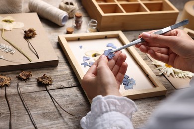 Photo of Woman putting dry flowers into picture frame at wooden table, closeup