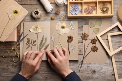 Photo of Woman putting dry flowers into notebook at wooden table, top view