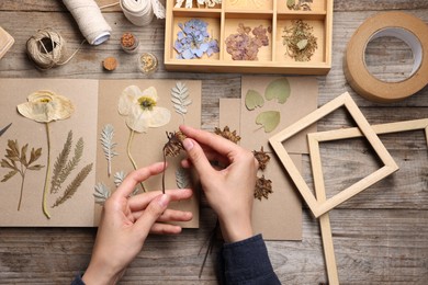 Photo of Woman putting dry flowers into notebook at wooden table, top view