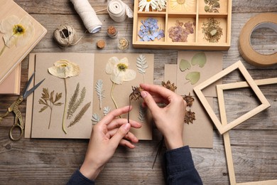 Photo of Woman putting dry flowers into notebook at wooden table, top view