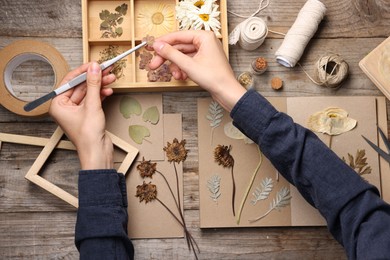 Photo of Woman putting dry flowers into notebook at wooden table, top view