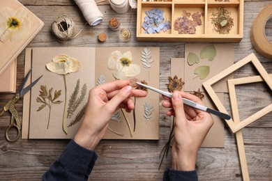Photo of Woman putting dry flowers into notebook at wooden table, top view