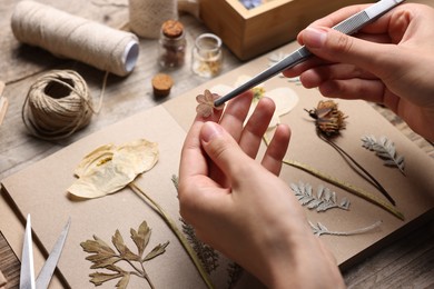 Photo of Woman putting dry flowers into notebook at wooden table, closeup