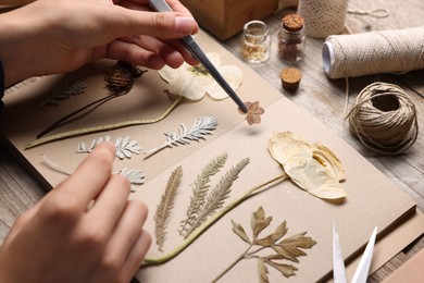 Photo of Woman putting dry flowers into notebook at wooden table, closeup