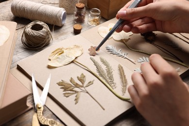Photo of Woman putting dry flowers into notebook at wooden table, closeup