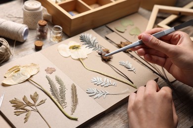 Photo of Woman putting dry flowers into notebook at wooden table, closeup