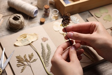 Photo of Woman putting dry flowers into notebook at wooden table, closeup