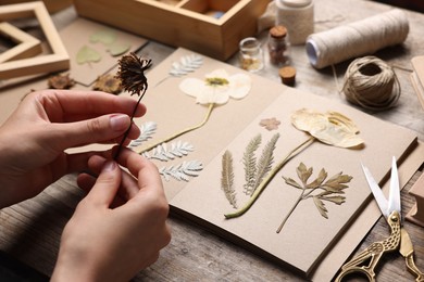 Photo of Woman putting dry flowers into notebook at wooden table, closeup