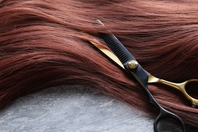 Photo of Auburn hair strand and hairdresser's scissors on grey background, closeup