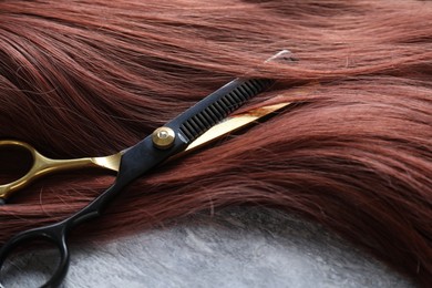 Photo of Auburn hair strand and hairdresser's scissors on grey background, closeup