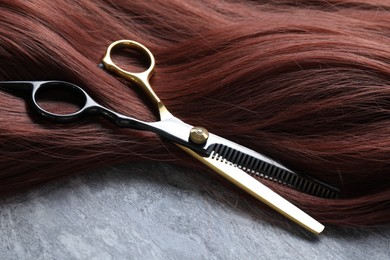 Photo of Auburn hair strand and hairdresser's scissors on grey background, closeup