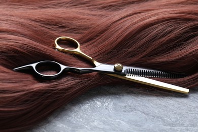 Photo of Auburn hair strand and hairdresser's scissors on grey background, closeup
