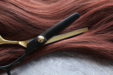 Photo of Auburn hair strand and hairdresser's scissors on grey background, closeup