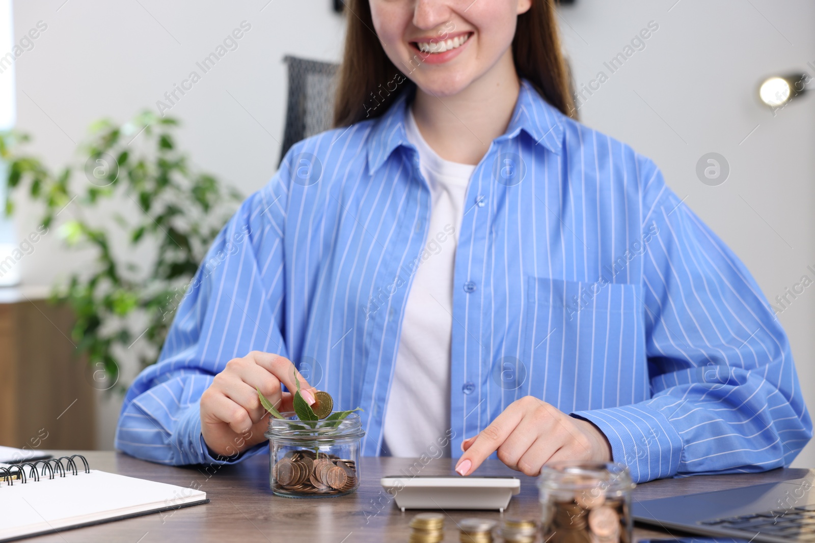 Photo of Woman putting coin into glass jar and using calculator at wooden table indoors, closeup