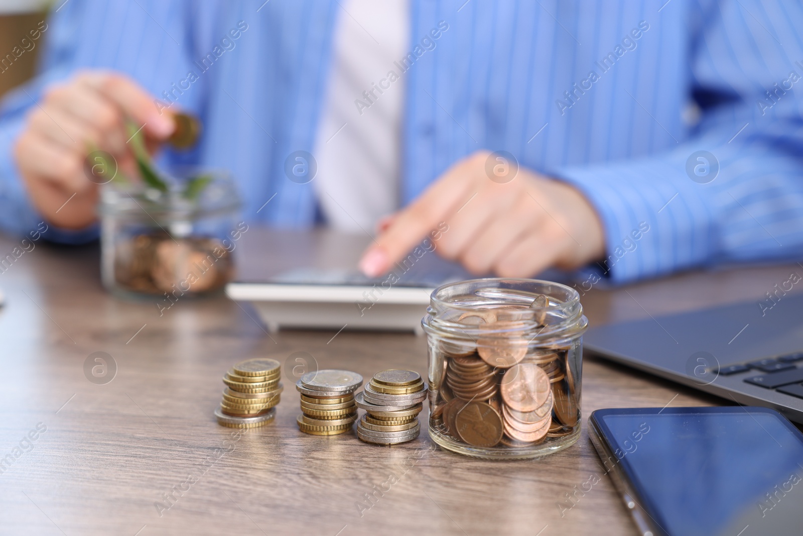 Photo of Woman putting coin into glass jar and using calculator at wooden table indoors, selective focus