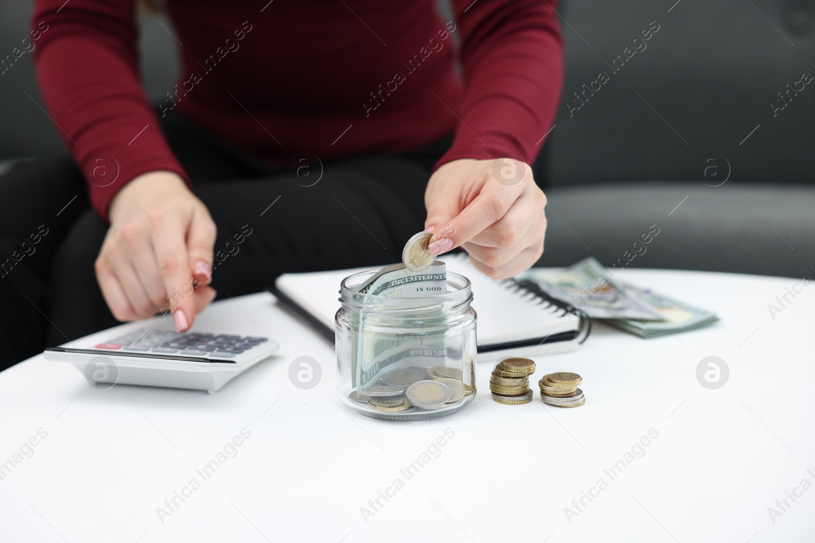Photo of Woman putting coin into glass jar and using calculator at table indoors, closeup