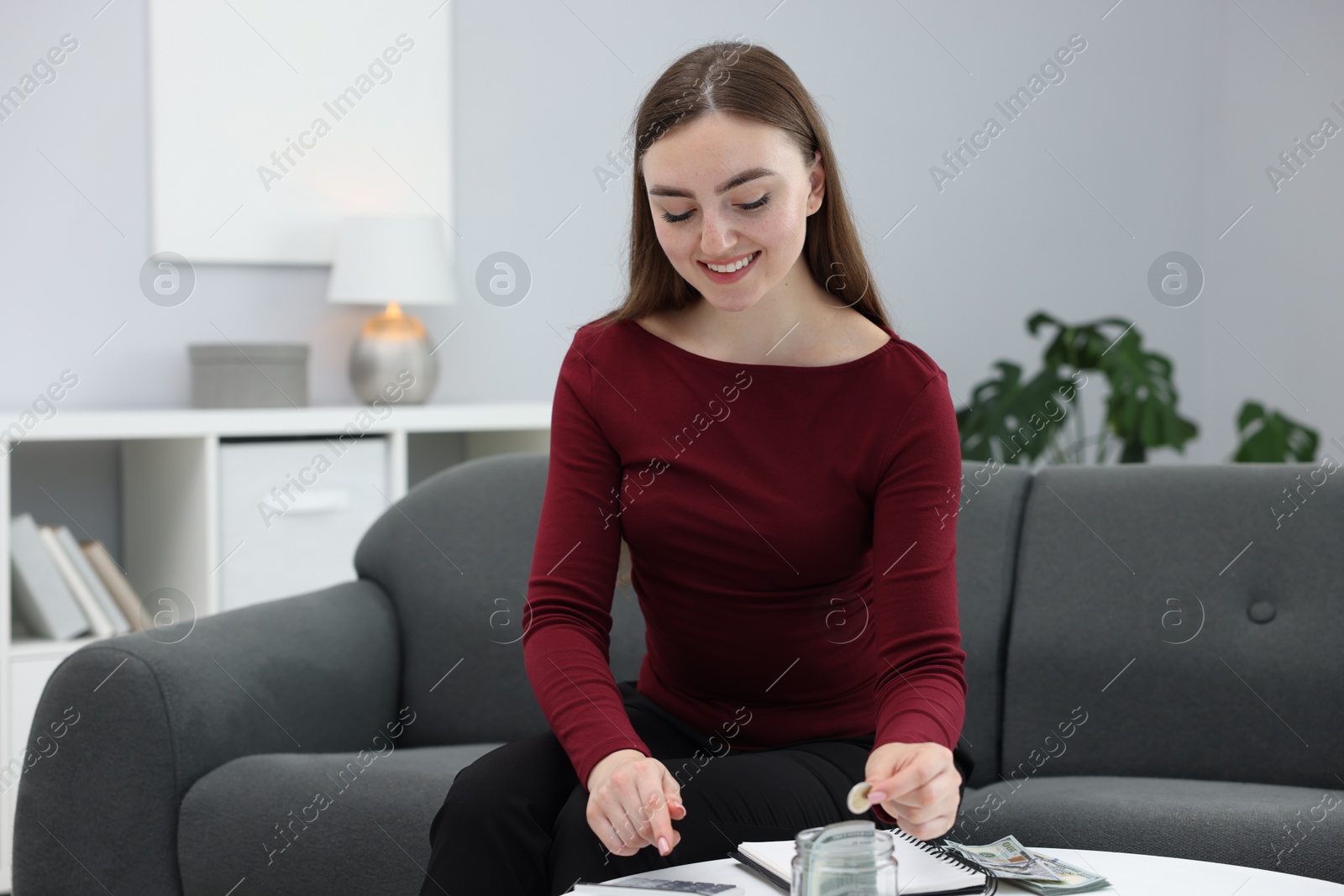 Photo of Woman putting coin into glass jar and using calculator indoors