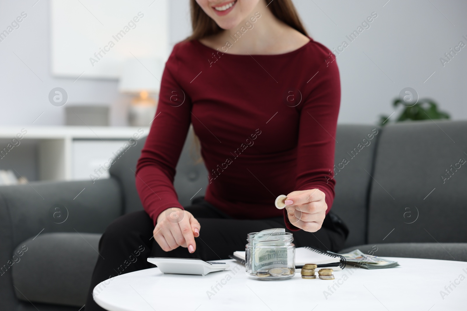 Photo of Woman putting coin into glass jar and using calculator at table indoors, closeup