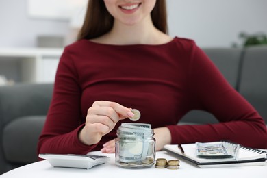 Photo of Woman putting coin into glass jar at table indoors, closeup