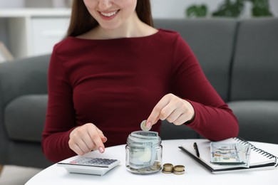 Photo of Woman putting coin into glass jar and using calculator at table indoors, closeup